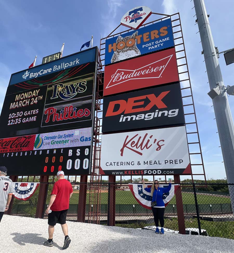 Wide angle view of Kelli in front of Kelli's Catering sign at BayCare park